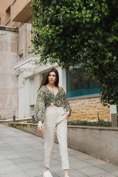 chic woman with long hair in trendy outfit with beige pants, cropped blouse and handbag with chain strap walking with hand in pocket near modern building and green tree on urban street in Istanbul
