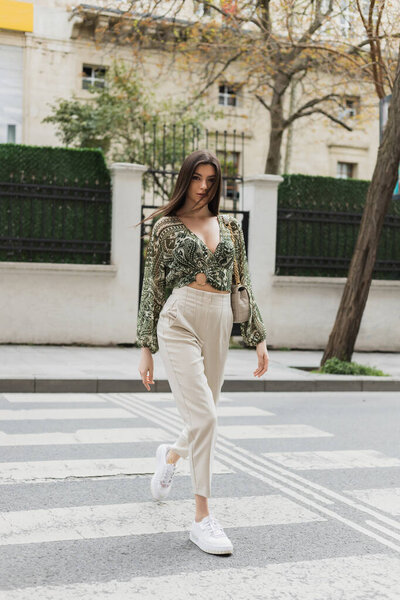 stylish woman with brunette long hair in trendy outfit with beige pants, cropped blouse and handbag with chain strap walking on crosswalk of urban street in Istanbul, blurred house on background 