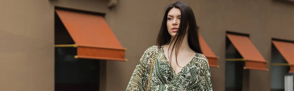 stylish woman with brunette long hair in trendy outfit with cropped blouse and handbag on chain strap standing near blurred building on urban street in Istanbul, banner 