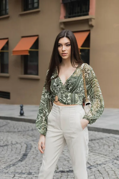 young woman with brunette long hair in beige pants, cropped blouse and handbag with chain strap standing with hand in pocket near blurred building on urban street in Istanbul