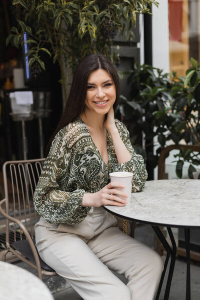 joyous young woman with long brunette hair and makeup sitting on chair near round bistro table and holding coffee in paper cup and smiling near blurred plants on terrace of cafe in Istanbul 
