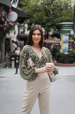 beautiful woman with long hair holding morning coffee in paper cup and newspaper while standing in trendy outfit with handbag and smiling on urban street near blurred flower shop in Istanbul  clipart