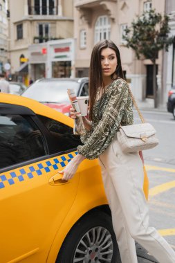 chic woman with long hair holding paper cup with coffee and newspaper while standing in trendy outfit with handbag on chain strap and opening door of yellow taxi on blurred urban street in Istanbul  clipart