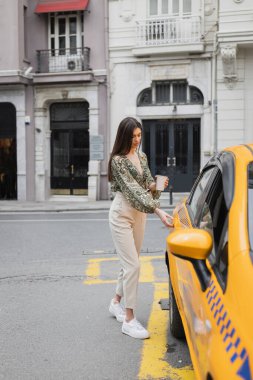 chic woman with long hair holding coffee in paper cup while standing in trendy outfit with handbag on chain strap and opening door of yellow cab on blurred urban street  clipart