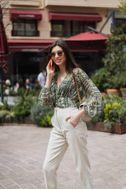 Uzun saçlı ve güneş gözlüklü neşeli genç kadın akıllı telefonla konuşurken gülümsüyor ve İstanbul 'da bulanık bina ve bitkilerin yanındaki zincir askıda el ve el çantasıyla ayakta duruyor. 