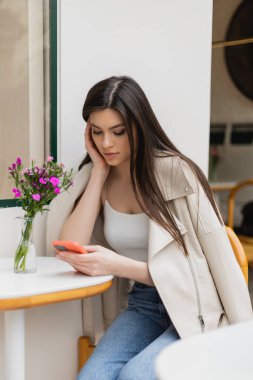 Uzun saçlı genç bir kadın, bistro masasının yanındaki vazoda çiçeklerle otururken ve akıllı telefon kullanırken İstanbul 'da terasta, terasta modaya uygun kıyafetler giyerken. 