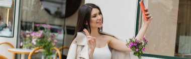 Uzun saçlı mutlu bir kadın selfie çekiyor, V işareti koyuyor, bej renkli deri ceketli modaya uygun kıyafetlerle oturuyor İstanbul 'da terasta bulanık çiçekler, afiş. 