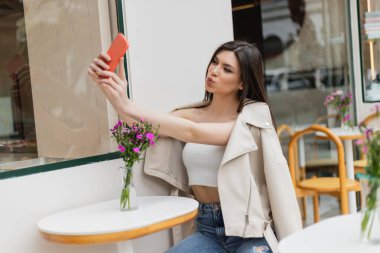 Uzun saçlı kadın, bistro masasının yanındaki sandalyede oturuyor vazoda çiçekler ve dudak büküyor, akıllı telefonlarda selfie çekiyor İstanbul 'da terasta son moda kıyafetler içinde poz verirken. 