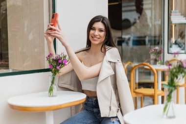 smiling woman with long hair sitting on chair near bistro table with flowers in vase and taking selfie on smartphone while posing in trendy clothes in cafe on terrace outdoors in Istanbul  clipart