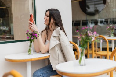 İstanbul 'da terasta, terasta modaya uygun giysiler içinde otururken bistro masasının yanında oturan uzun saçlı mutlu kadın vazoda çiçekler ve akıllı telefonla mesajlaşıyor. 