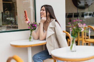 İstanbul 'da terasta bej ceketli ve modaya uygun kıyafetler içinde otururken, bistro masasının yanındaki sandalyede oturan uzun saçlı, vazoda çiçekli ve akıllı telefonuyla mesajlaşan mutlu bir kadın. 