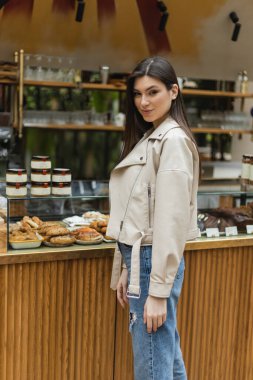 brunette young woman with long hair in beige leather jacket and denim jeans looking at camera while standing near cake display with tasty pastry and jars of jam in modern bakery shop in Istanbul  clipart