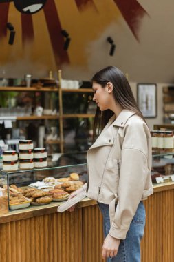 young woman with brunette long hair in beige leather jacket and denim jeans standing near cake display with delicious pastry and jars of jam in modern bakery shop in Istanbul  clipart
