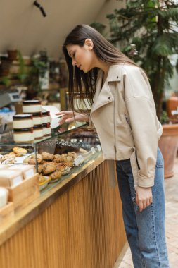 brunette young woman with long hair in beige leather jacket and denim jeans standing near cake display and choosing pastry near jars of jam in modern bakery shop in Istanbul  clipart