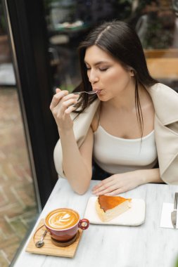 young woman with long hair eating cheesecake next to cup of cappuccino on bistro table while sitting in leather jacket next to window inside of modern cafe in Istanbul  clipart