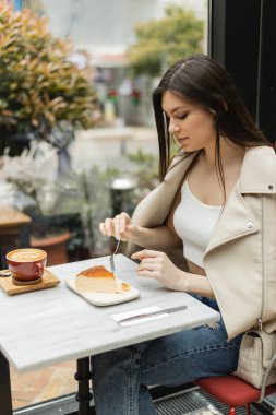 young  brunette woman holding fork near cheesecake next to cup of cappuccino with coffee art on bistro table while sitting in leather jacket near window inside of modern cafe in Istanbul  clipart