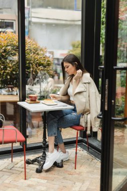 full length of young woman with long hair holding fork near cheesecake and cup of cappuccino on bistro table while sitting in leather jacket next window inside of modern cafe in Istanbul  clipart