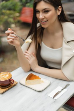 smiling woman with long hair holding fork with cheesecake next to cup of cappuccino with coffee art on bistro table while sitting inside of modern cafe in Istanbul  clipart