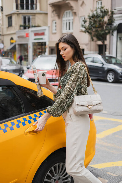 pretty woman with long hair holding paper cup with coffee and newspaper while standing in trendy outfit with handbag on chain strap and opening door of yellow taxi on blurred urban street in Istanbul 