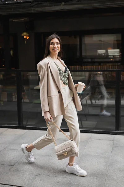 smiling woman with long hair walking in blazer with beige pants, cropped blouse and handbag on chain strap and holding paper cup with coffee near outdoor cafe on urban street in Istanbul