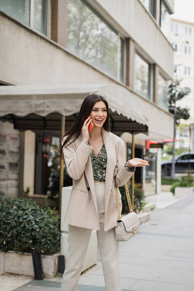 happy young woman with brunette long hair and makeup smiling while talking on smartphone and standing in trendy outfit with handbag on chain strap near blurred fancy restaurant in Istanbul 