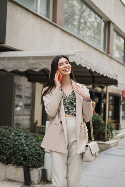 smiling young woman with brunette long hair and makeup smiling while talking on smartphone and standing in trendy outfit with handbag on chain strap near blurred restaurant in Istanbul 