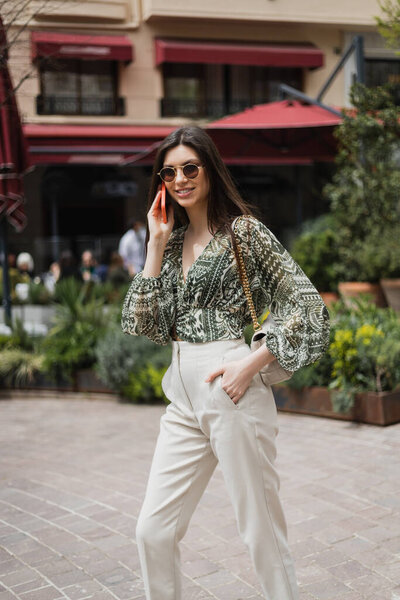 cheerful young woman with long hair and sunglasses smiling while talking on smartphone and standing with hand in pocket and handbag on chain strap near blurred building and plants in Istanbul 
