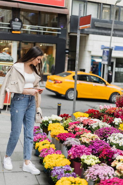 brunette woman with long hair standing in beige leather jacket, denim jeans and handbag with chain strap holding smartphone near flowers next to blurred street in Istanbul, vendor