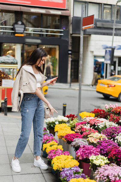 brunette woman with long hair standing in beige leather jacket and denim jeans while holding smartphone and pointing at bouquets of flowers next to blurred car on street in Istanbul, vendor