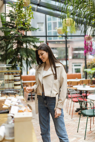 brunette young woman with long hair in beige leather jacket and denim jeans standing near cake display with pastry and jars of jam in modern bakery shop in Istanbul 