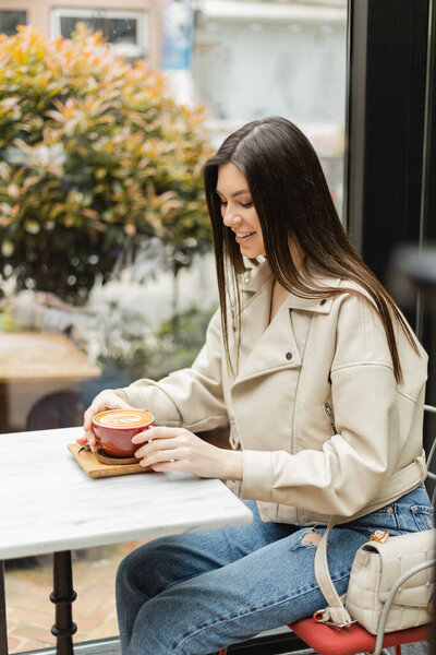 happy brunette woman in leather jacket sitting on chair next to window and bistro table while holding cup of cappuccino with coffee art inside of modern cafe in Istanbul 