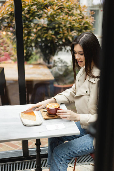 happy woman with long hair sitting in leather jacket next to window and bistro table while holding cup of cappuccino with coffee art and looking at cheesecake inside of modern cafe in Istanbul 