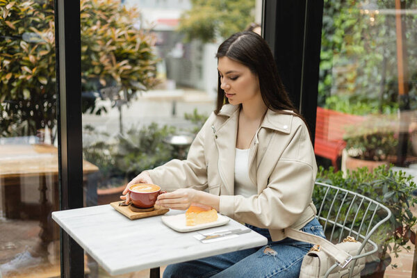brunette woman with long hair sitting in leather jacket next to window and bistro table while holding cup of cappuccino with coffee art near cheesecake inside of modern cafe in Istanbul 