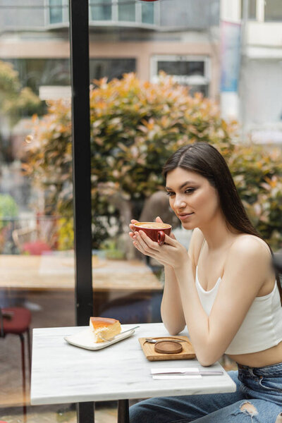 happy woman with long hair sitting next to window and holding cup of cappuccino with coffee art near cheesecake on plate of bistro table inside of modern cafe in Istanbul 