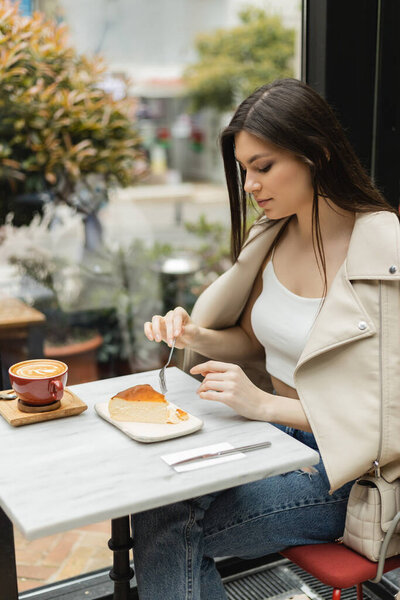 young  brunette woman holding fork near cheesecake next to cup of cappuccino with coffee art on bistro table while sitting in leather jacket near window inside of modern cafe in Istanbul 