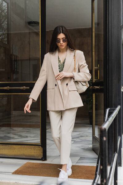 brunette and young woman with long hair walking out of modern building in stylish outfit and sunglasses while holding handbag with chain strap on street in Istanbul, entrance door 