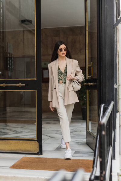 brunette and young woman with long hair in stylish outfit and sunglasses walking out of modern building while holding handbag with chain strap on street in Istanbul, entrance door 