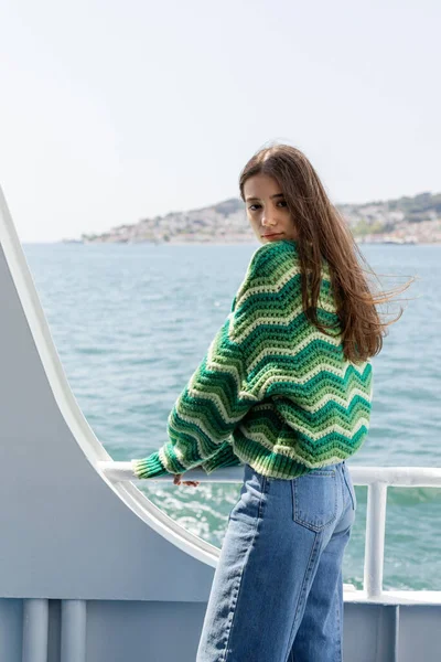 Mujer joven en suéter de punto y jeans mirando a la cámara mientras está de pie en el ferry con las islas de la princesa en el fondo - foto de stock