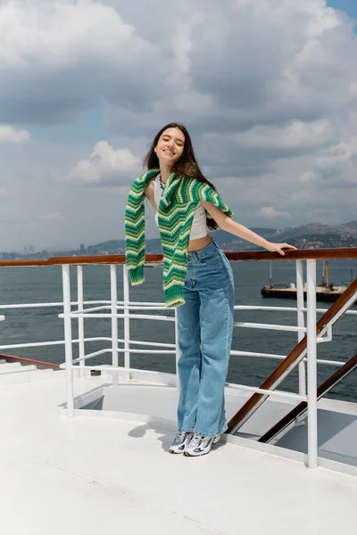 Happy young tourist in sweater and jeans looking at camera near railing on ferry boat in Turkey — Stock Photo