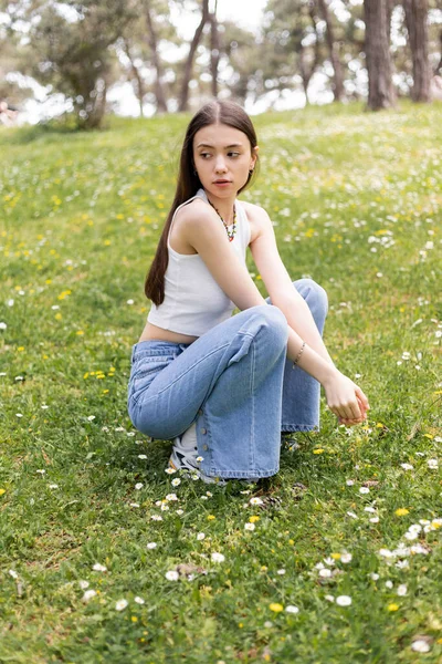 Jeune femme en haut et jeans assis sur la pelouse avec des marguerites le jour — Photo de stock