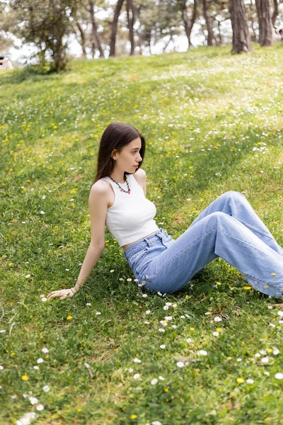 Vue latérale de la jeune femme haut et jeans assis sur la prairie avec des fleurs en été — Photo de stock
