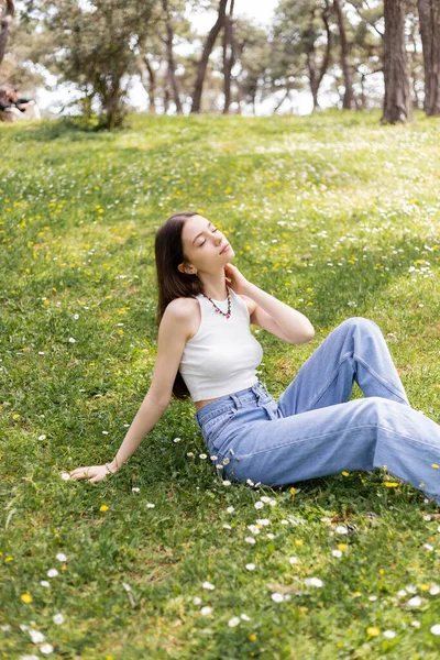 Молодая женщина в верхней части и джинсы касаются шеи, сидя на газоне с цветами в парке — стоковое фото