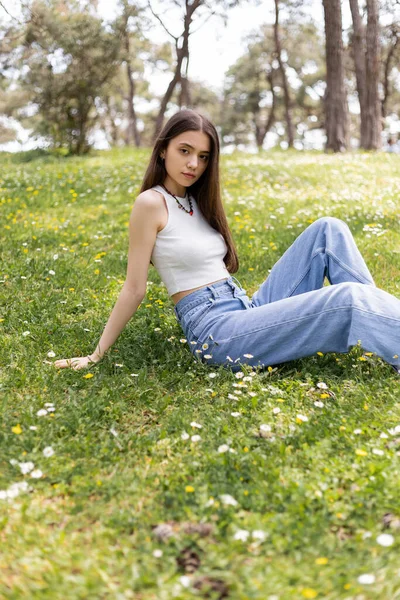Jovem morena em cima e jeans olhando para a câmera enquanto sentado no gramado com flores no parque — Fotografia de Stock