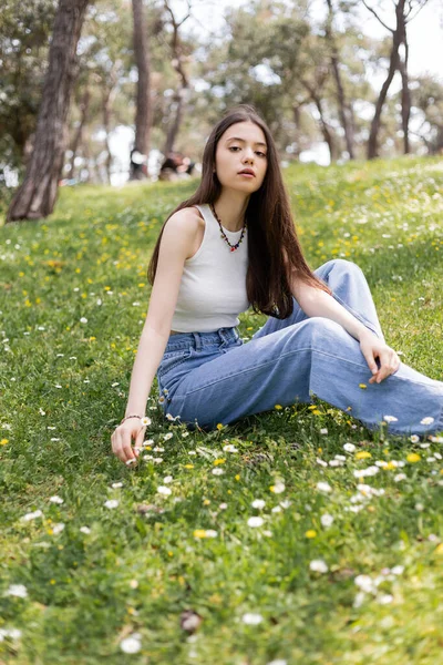 Jovem mulher em roupas casuais olhando para a câmera enquanto sentado no prado com flores no parque — Fotografia de Stock