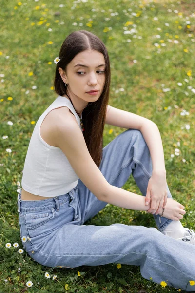 Молодая женщина наверху и джинсы, сидящие на газоне с цветами маргаритки в парке — стоковое фото