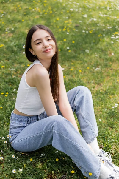 Overjoyed woman in top and jeans looking at camera while sitting on lawn with flowers in park — Stock Photo