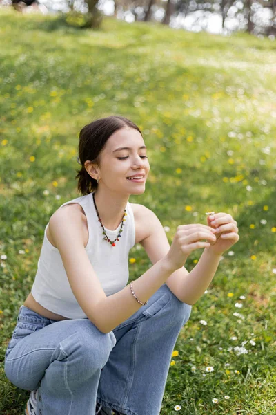 Усміхнена молода жінка зверху і джинси, що тримають ромашкову квітку на газоні в парку — стокове фото