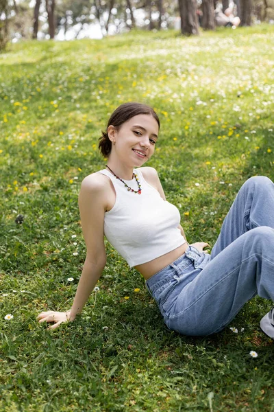 Сверхрадостная молодая женщина в джинсах и сверху смотрит в камеру, сидя на газоне в парке — стоковое фото