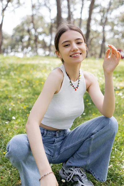 Mujer joven sonriente en la parte superior y jeans mirando a la cámara en el prado borroso con flores en el parque - foto de stock