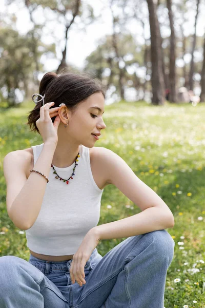 Vista lateral de la joven mujer poniendo la flor de margarita en el pelo mientras está sentado en el césped en el parque - foto de stock